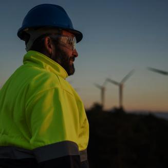 A person wearing a bright yellow high‑visibility jacket, dark safety gloves, and a blue hard hat stands outdoors at dusk, holding a closed laptop at their side. In the distance, several wind turbines rise above a darkened landscape under a fading sky