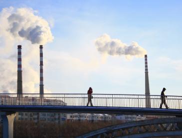People walk across a bridge as industrial smokestacks release smoke into a blue sky.